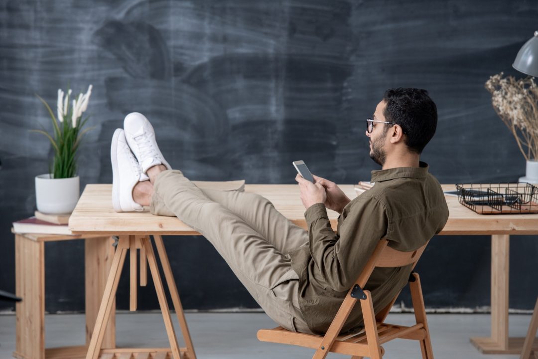 young employee in casualwear keeping legs on table while scrolling in smartphone_marcommsinc